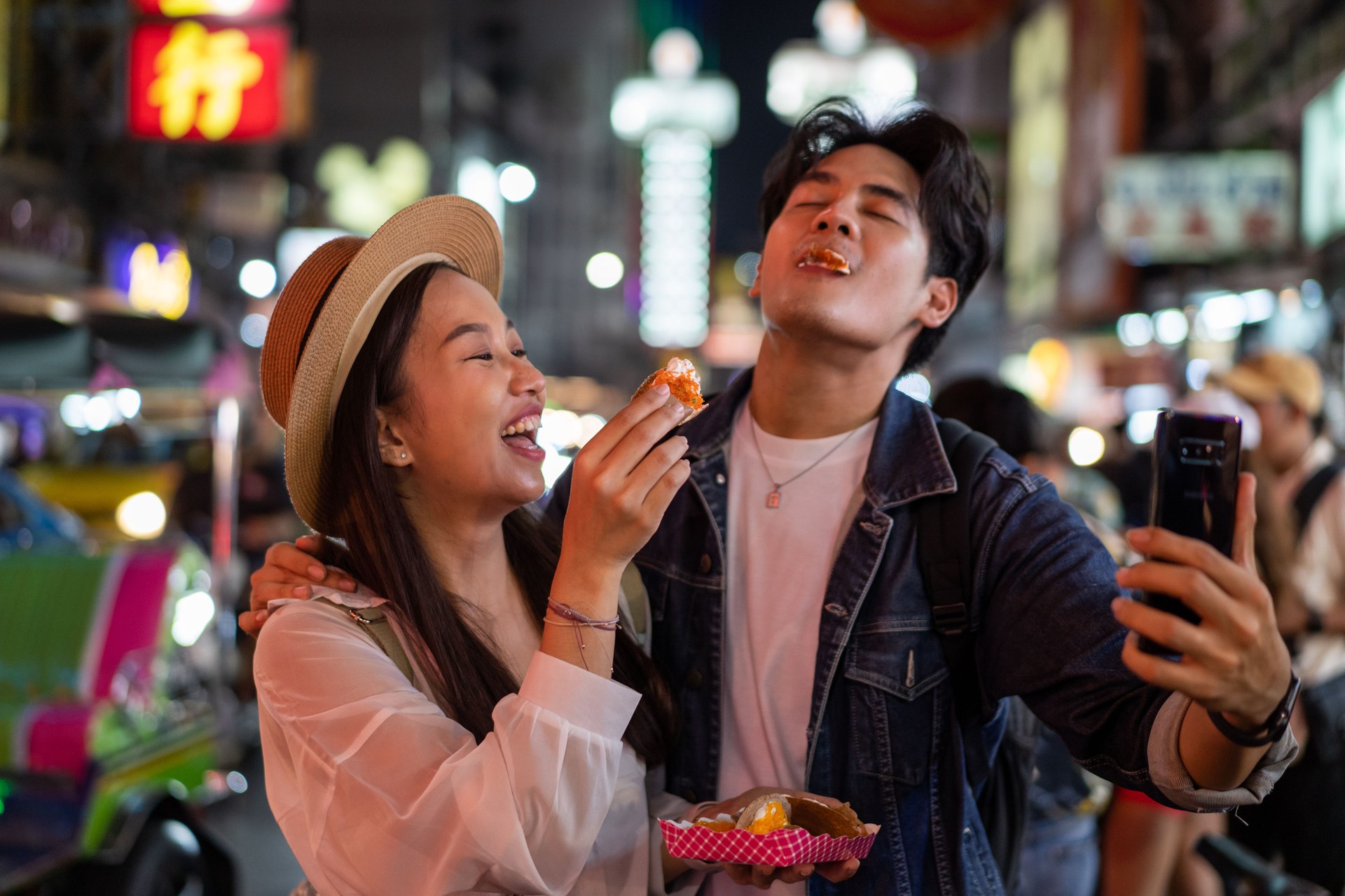 Asian couple tourist backpacker doing selfie and recording a video blog while eating food from street stall in night market with crowd of people at Yaowarat road, Bangkok.