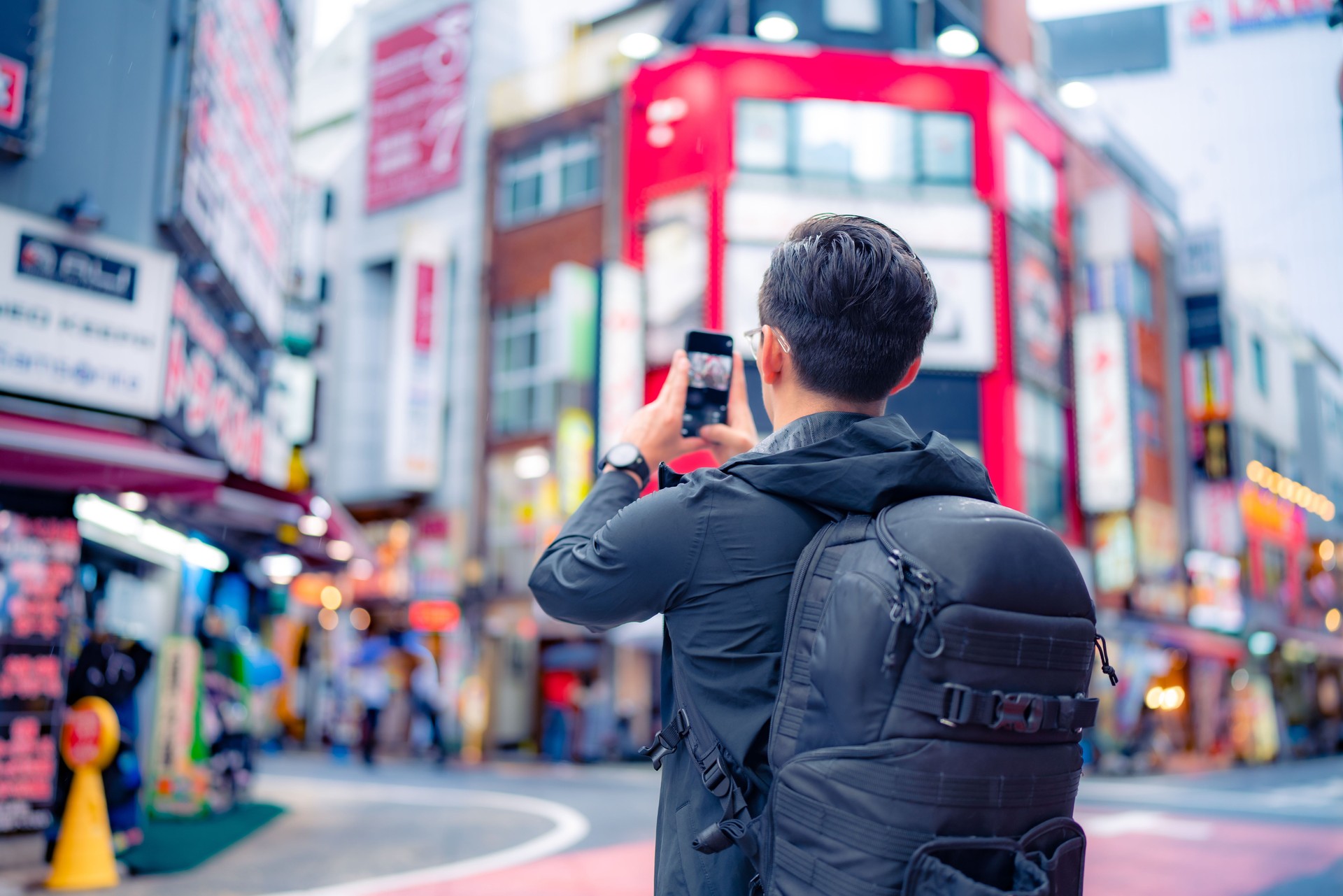Back view of unrecognizable Hispanic male tourist with backpack taking picture on smartphone in bright Shinjuku neighborhood street of Tokyo city, Japan